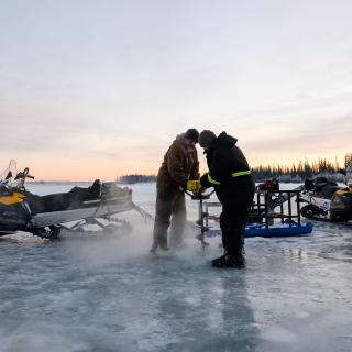 Two people using an ice auger to drill through a frozen lake to ice fish. Sunset and snowmachines in background.