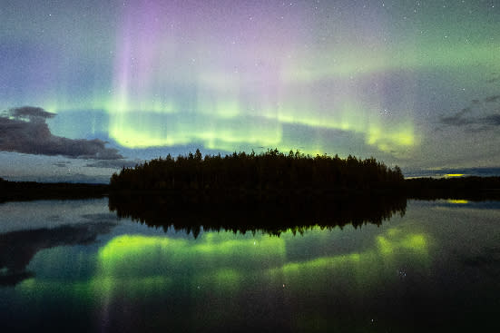 Green and purple northern lights above a dark forest island, with colorful reflections shimmering on lake