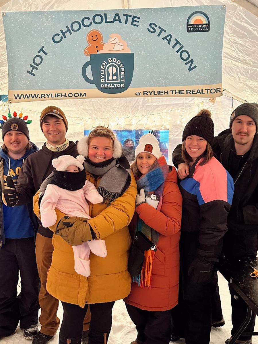 A group of people in winter clothing, standing and smiling underneath a sign that says, "Hot Chocolate Station."