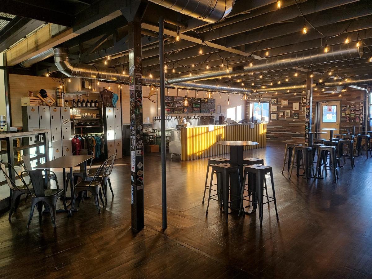 Interior of Lat 65 taproom featuring industrial-style decor with exposed beams, string lights, metal chairs and stools, a polished wood floor, and a warmly lit bar.
