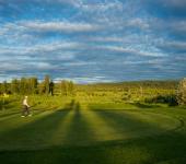 Golf course with vibrant grass, long shadows, and cloudy sky. A man is walking through the center of the image.