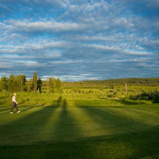 Golf course with vibrant grass, long shadows, and cloudy sky. A man is walking through the center of the image.