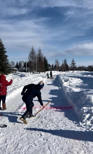 A person preparing to slide a wooden stick across the ice in the Snow Snake event at the Dena Games, while spectators watch from the sidelines under a partly cloudy sky.