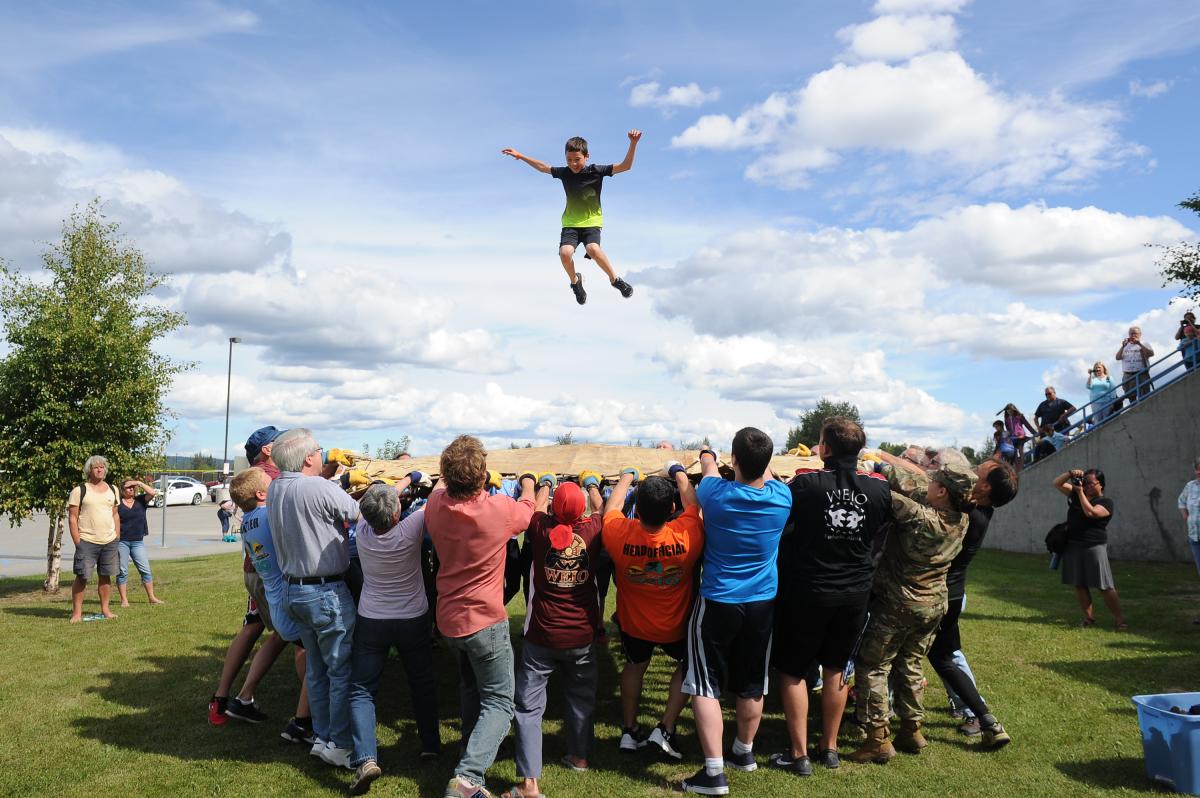 A group of people outdoors tossing a child into the air as part of the Nalukataq (blanket toss) event on a sunny day with blue sky and scattered clouds.