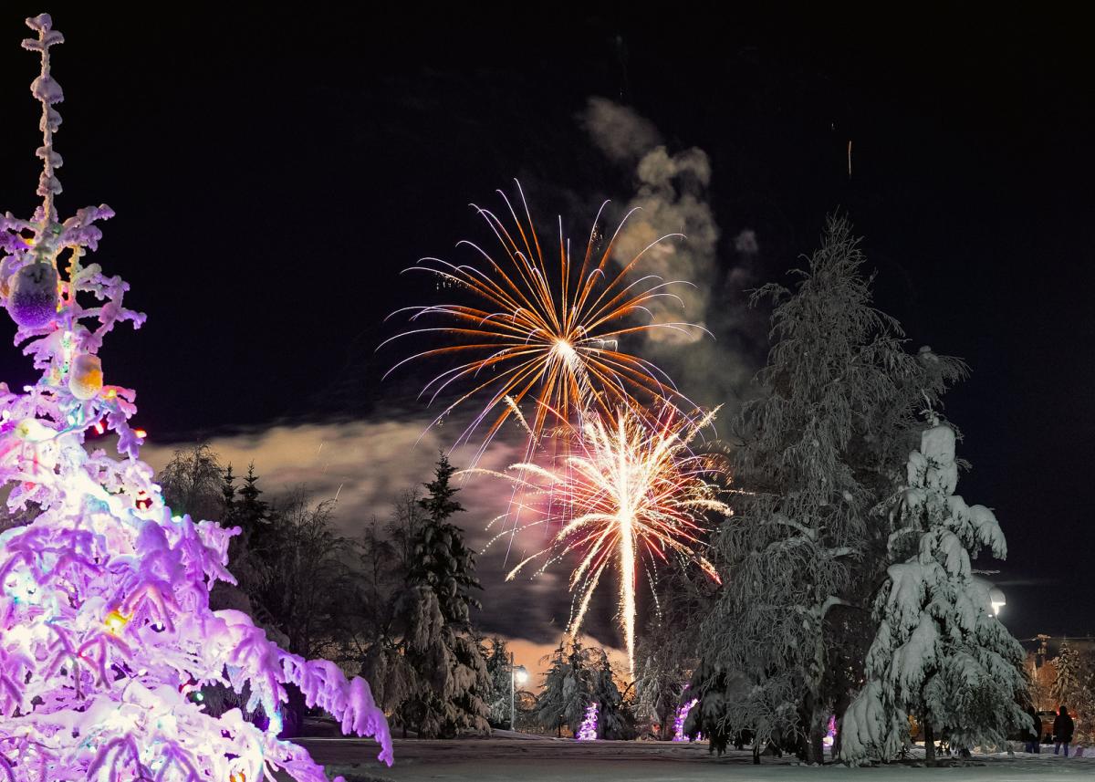 Colorful fireworks lighting up the night sky above snow-covered trees, with a glowing Christmas tree in the foreground.